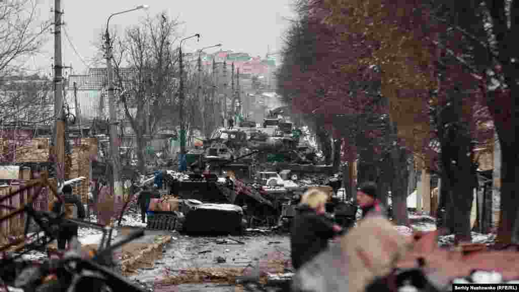 Мірныя жыхары глядзяць на зьнішчаную расейскую бранятанкавую калёну ў Бучы, непадалёк ад Кіева, 1 сакавіка 2022. Горад быў канчаткова захоплены расейскімі войскамі празь некалькі дзён пасьля таго, як зрабілі гэты фатаздымак, але яны адступілі менш чым празь месяц. Пасьля адступленьня расейскіх войскаў у Бучы пачалі фіксаваць доказы ваенных злачынстваў Расеі, якія выклікалі міжнароднае асуджэньне.